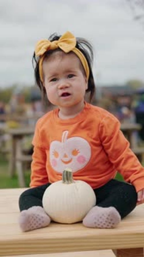 Adorable Baby in Orange Pumpkin Sweater with Yellow Headband Sitting Outdoors Holding a Small White