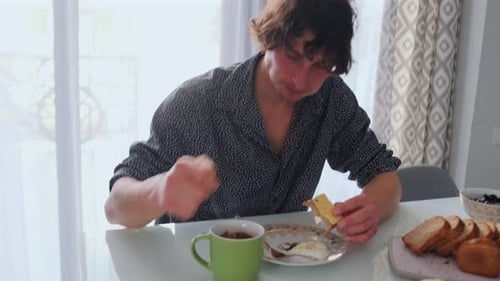 Young Man Drinking Coffee at Breakfast Sunlit Kitchen Table with Croissant Toast Butter Boiled Egg