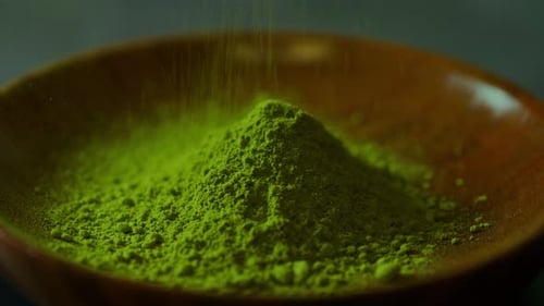 A vibrant heap of finely ground matcha green tea powder sits in a wooden bowl as more powder gently