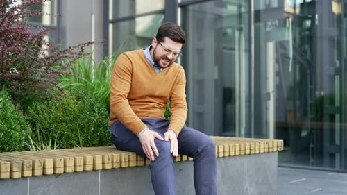Businessman suffers from painful severe knee joint pain sitting on bench on street near office