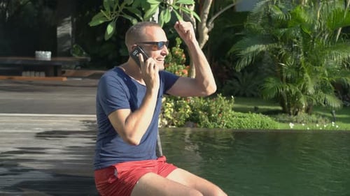 Handsome man relaxes by swimming pool chatting on cellphone during summer vacation