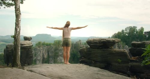 Woman with long hair stands on a rocky cliff, arms outstretched, embracing the breathtaking view of