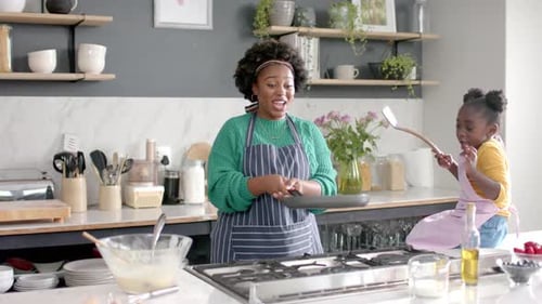 Happy african american mother and daughter frying pancakes in kitchen, slow motion
