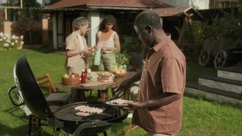 African American Dad Placing Sausages on Barbecue Grill and Chatting to Family