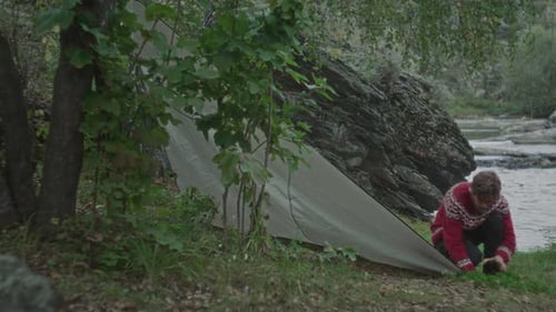 Man Anchoring Tent Edge to Ground near River and Rocks in Forest