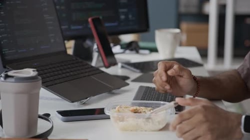 Focused Indian Employee Eating Lunch at Office Desk While Coding on Computer