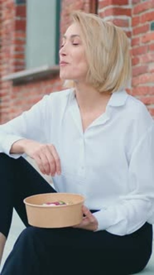 Woman Eating Healthy Salad with Olive From Plastic Container Sitting on Stairs in City Street