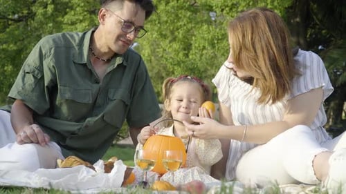 A young happy family at the picnic in the city park. A little girl putting tangerine in the hallowee