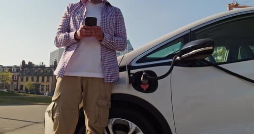 Man Uses Smartphone Next to Charging Electric Vehicle