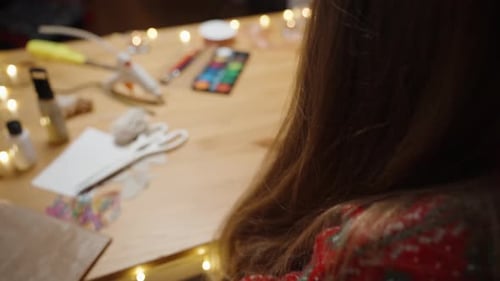 A Woman in a Red Sweater Cuts Paper for a Craft Project at a Table with Art Supplies and Christmas D