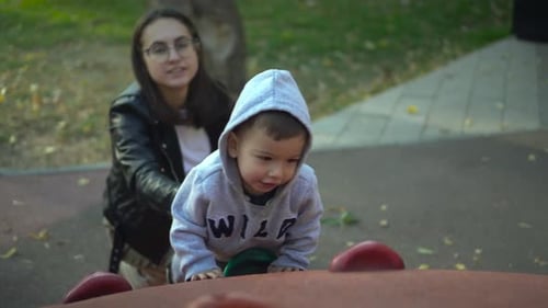 Mom Helps Her Son Climb the Slide A Boy on the Playground Climbs Up a Hill A Child in a Gray Hoodie