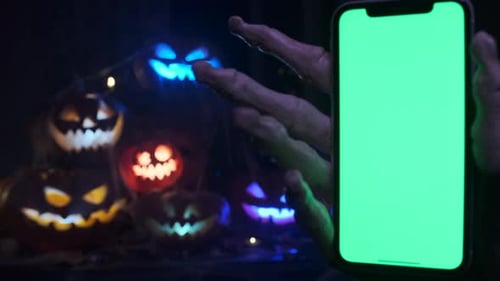 Creepy hand with phone chroma key screen against backdrop of creepy jack-o'-lanterns