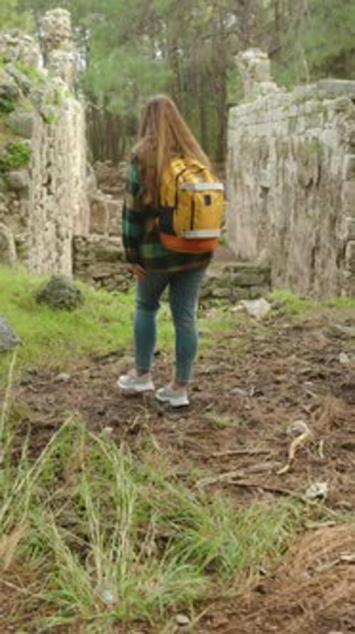 Vertical video. A female tourist with a backpack walks through the forest among the ruins.