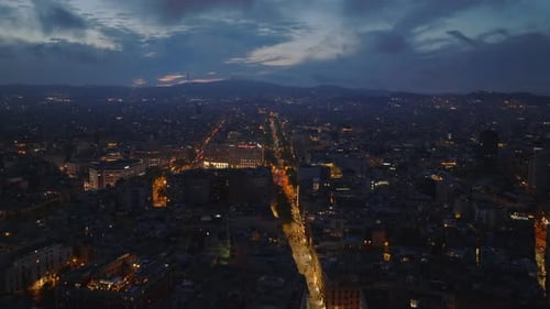 Fly Above Night City Cityscape with Illuminated Streets and Hills Against Colour Sky in Background