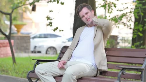 Man Sits on Bench in Park With Neck Pain