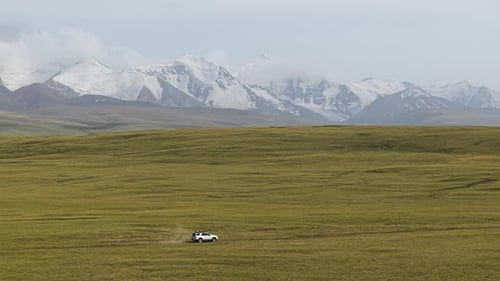 Vehicle Driving Through A Vast, Open Meadow With Snow-capped Mountains In The Background In