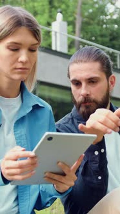 Two Colleagues Sitting on the Grass Outdoors Collaborating with a Laptop and a Tablet The Man