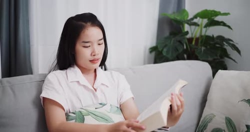 Woman relaxing on sofa reading a book in living room at home.