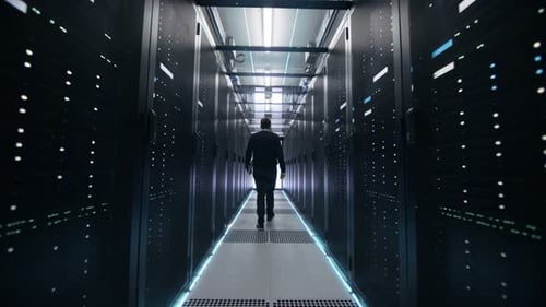 Foto siguiente de un ingeniero de TI recorriendo el pasillo del centro de datos con filas de servidores en rack.
Abre