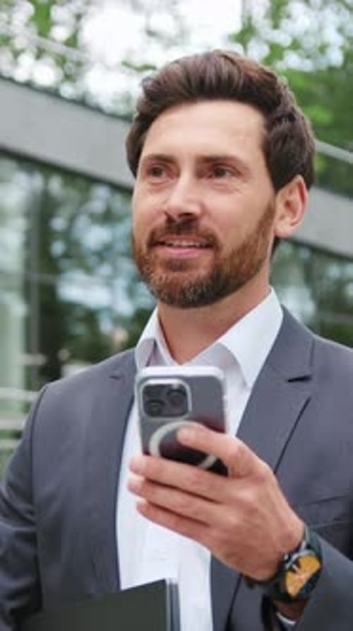 Confident Businessman in a Formal Suit Holding a Laptop and Using His Smartphone for Online