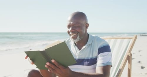 Happy senior african american man sitting on deck chair and reading book at beach, in slow motion