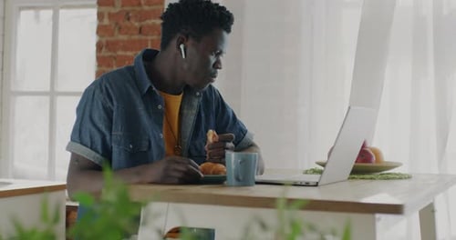 Happy African American Guy Eating Pastry Drinking Tea and Watching Funny Video on Laptop in Kitchen