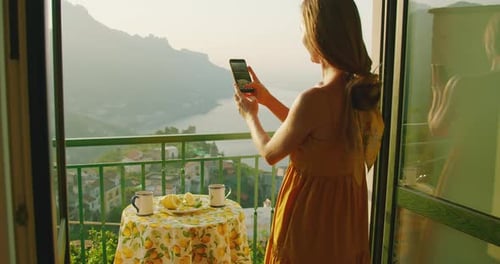 A woman on a balcony overlooking the Amalfi Coast, Italy. Tourist films breakfast on her smartphone
