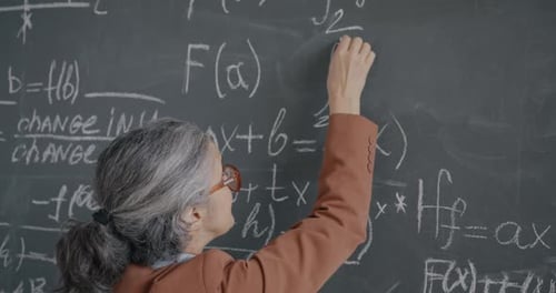 High School Teacher Senior Woman Writing Formula on Chalkboard and Speaking Teaching Algebra in