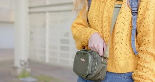 Female student pausing on sidewalk, unzipping bag pocket, grabbing smartphone checking notification