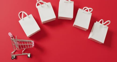 Five white gift bags and shopping trolley on red background with copy space