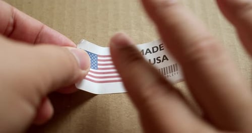 Hands applying MADE IN USA flag label on a shipping box with product premium quality barcode. Manufa