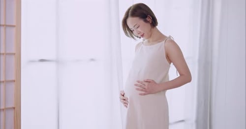 Happy Asian pregnant woman lovingly touching her unborn baby while standing by the window at home.