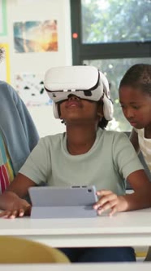 Starting VR, child in green T-shirt tilting head and holding tablet at desk, teacher monitoring