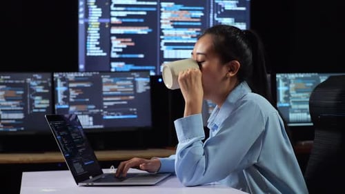 Seitenansicht einer asiatischen Programmiererin, die Kaffee trinkt, während sie Code an einem Laptop schreibt