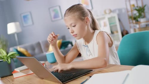 Smart Little Girl Does Homework in Her Living Room. She's Sitting at Her Desk, Uses Laptop and Writ