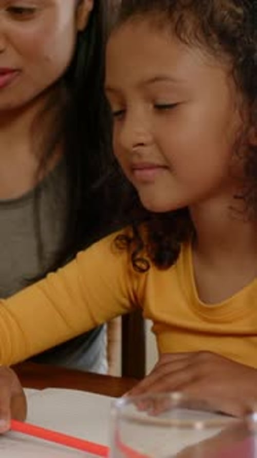 Vertical video: Leaning yellow kid tracing red pencil on paper at table mom guiding hand learning
