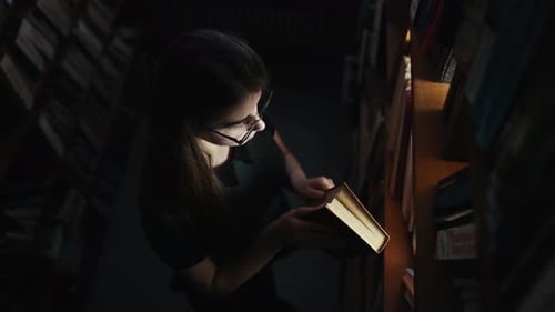Mädchen mit Brille auf der Suche nach dem Buch in einem dunklen Raum mit Lichtstrahl kommt durch die Regale