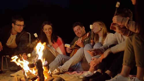 Friends roasting marshmallows singing and playing guitar at a campfire on the beach