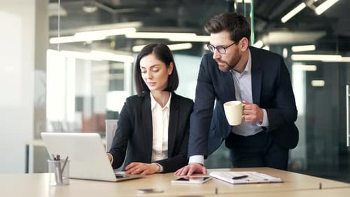 Colleagues collaborating in office setting discussing work on laptop. Man holding coffee cup points