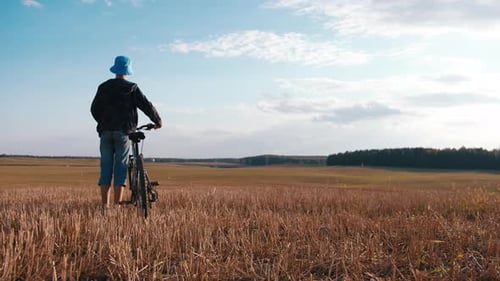 Teen Boy with a Bicycle Walks Along a Mowed Field in the Evening at Summer Static Shot