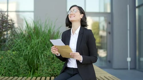 Joyful excited young businesswoman reading letter with great news sitting on a bench on the street
