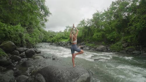 Slender female practicing Tree Pose outdoor next to jungle river, Vrksasana