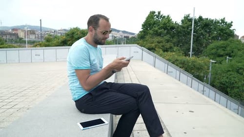 Young man texting on smartphone sits relaxing on city bench in sunlight