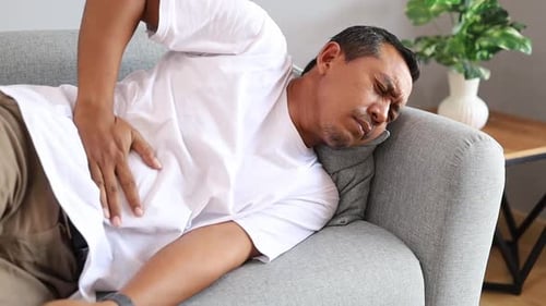 Asiatischer Mann hat Bauchschmerzen, während er drinnen auf einer Couch liegt