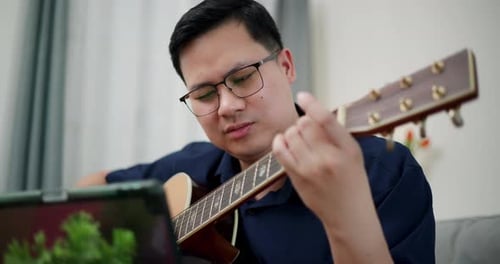 Homme asiatique détendu jouant joyeusement de la musique avec une guitare sur le canapé à la maison.