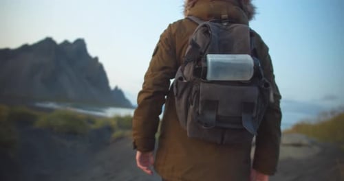 Hiker stands with his backpack on a black dune on the beach in front of a rocky mountain landscape