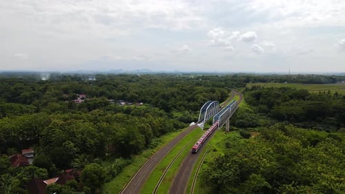 Aerial follow shot of train crossing river bridge in rural area of Indonesia during cloudy day
