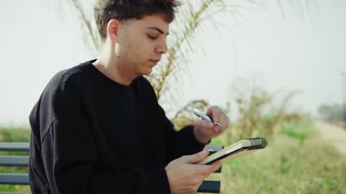 Young Student Writing Notes With A Pen While On The Park Bench