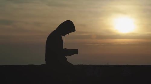A Young Man in a Hood at Sunset Reads a Book
