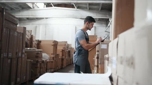 Young male worker in uniform in warehouse counting products and writing in notepad